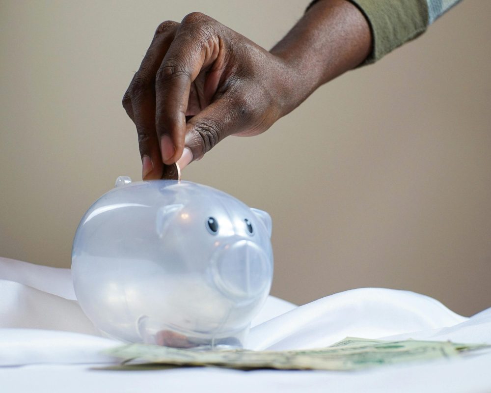 Close-up of a person's hand placing coins into a transparent piggy bank to save money.